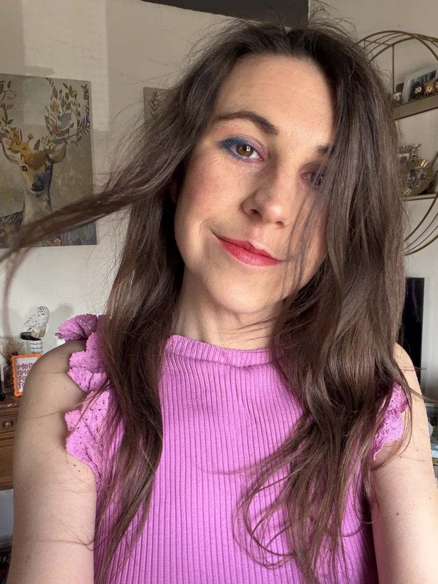 Camille Sauton chez elle : cheveux longs châtains, haut rose, devant une peinture de cerf et une étagère. Lumière douce de jour.